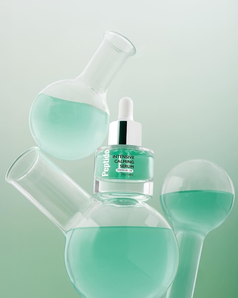 A high-end product hero shot featuring an intensive calming serum bottle positioned among lab glassware filled with teal liquid, conveying a clinical yet clean aesthetic.